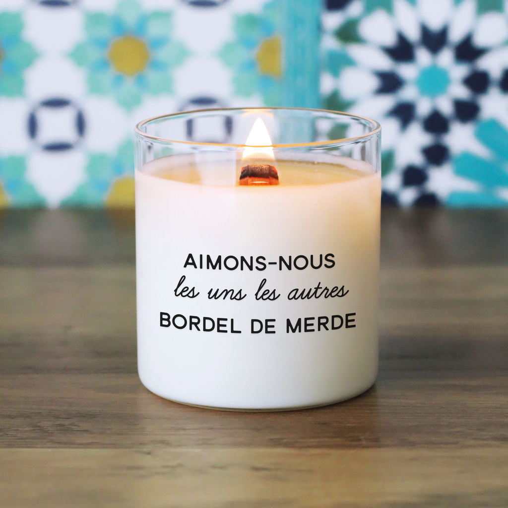 Bougie humoristique avec le message "Aimons-nous les uns les autres, bordel de merde", allumée sur fond à motifs colorés, parfaite pour un cadeau engagé et décalé.