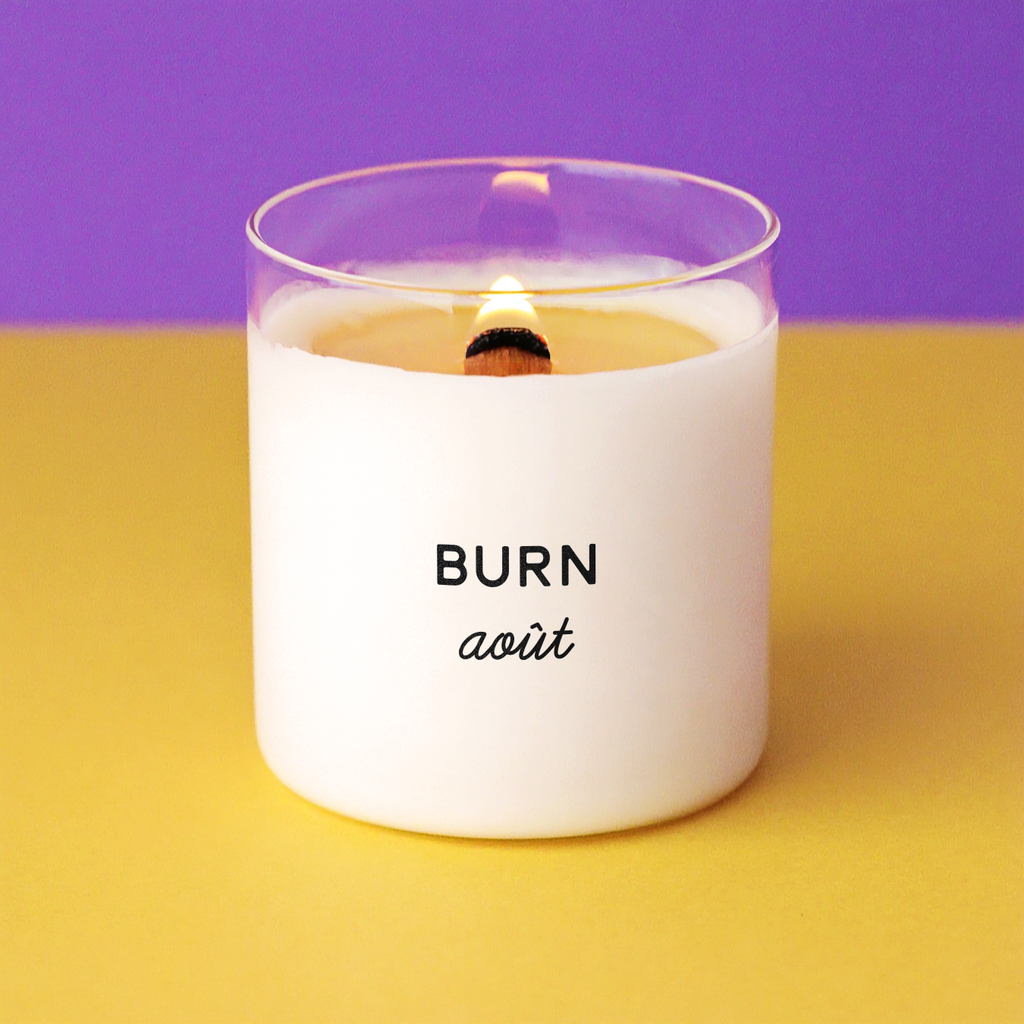 Bougie allumée avec l’inscription « BURN août », posée sur un fond jaune vif avec un mur violet en arrière-plan, parfaite comme idée cadeau humoristique pour les vacanciers ou les burn-outés du mois d’août.