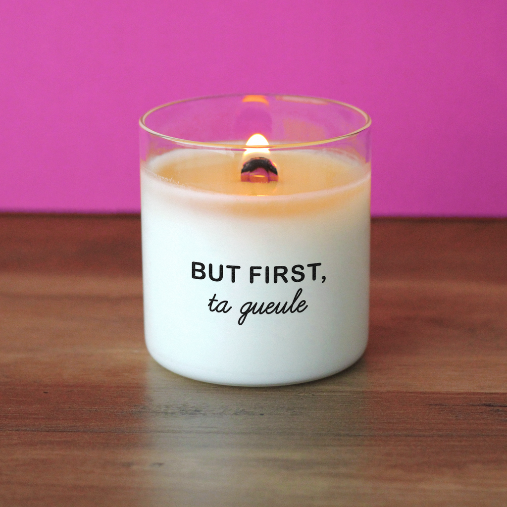 Bougie allumée avec l’inscription "But first, ta gueule", posée sur une table en bois avec fond rose fuchsia, parfaite comme cadeau drôle, insolent ou message décalé à offrir à un(e) ami(e).