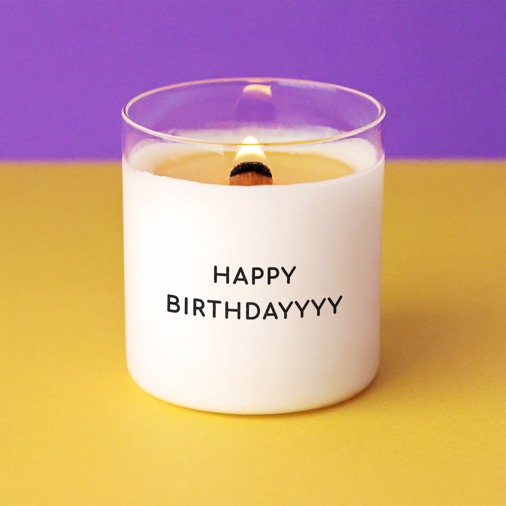 Bougie d’anniversaire avec le message HAPPY BIRTHDAYYYY, mèche en bois allumée, sur fond violet et jaune – idée cadeau humoristique pour anniversaire.