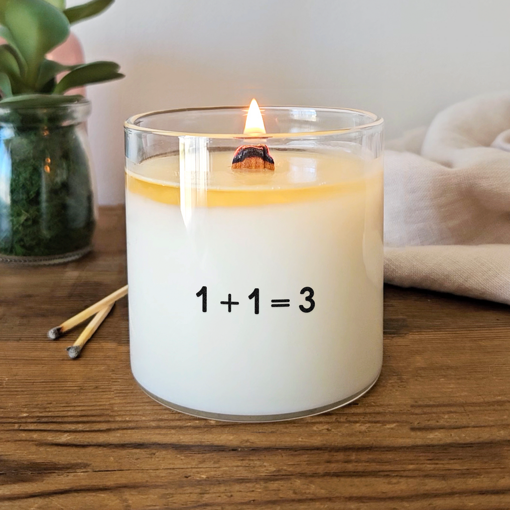 Bougie blanche avec l’inscription « 1 + 1 = 3 » en noir, sur une étiquette transparente. Idée cadeau ou annonce originale et tendre de grossesse.