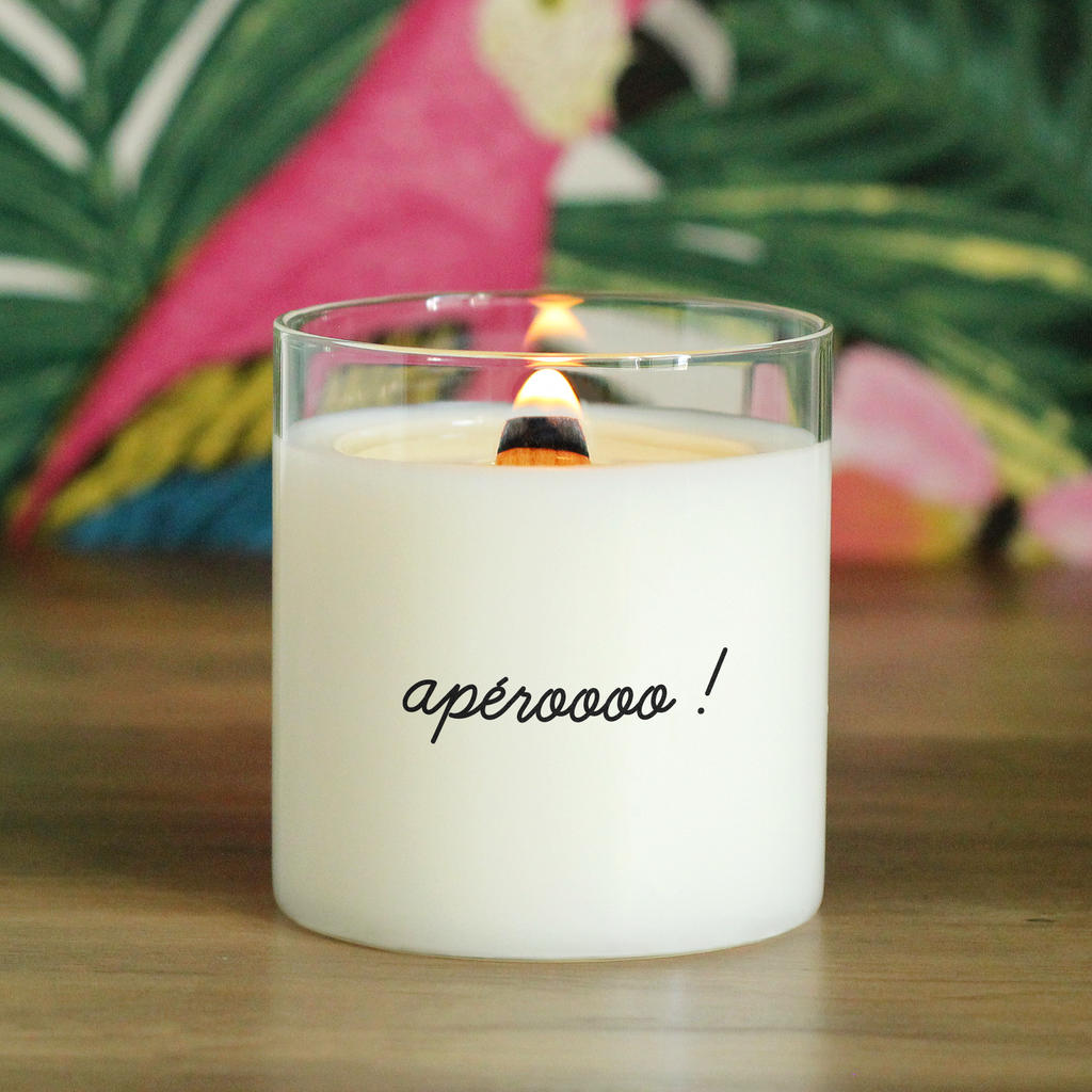 Bougie allumée avec l’inscription « apéroooo ! », posée sur une table en bois avec en fond un motif tropical coloré, parfaite pour l’été ou en cadeau humoristique entre amis.