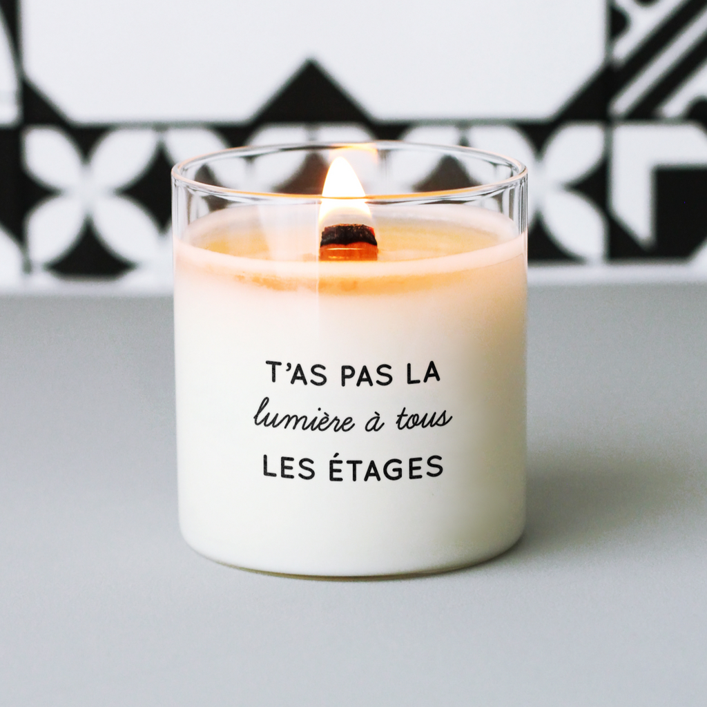Bougie blanche allumée avec le message "T’as pas la lumière à tous les étages", posée sur fond gris avec motif noir et blanc, parfaite comme cadeau humoristique et original.
